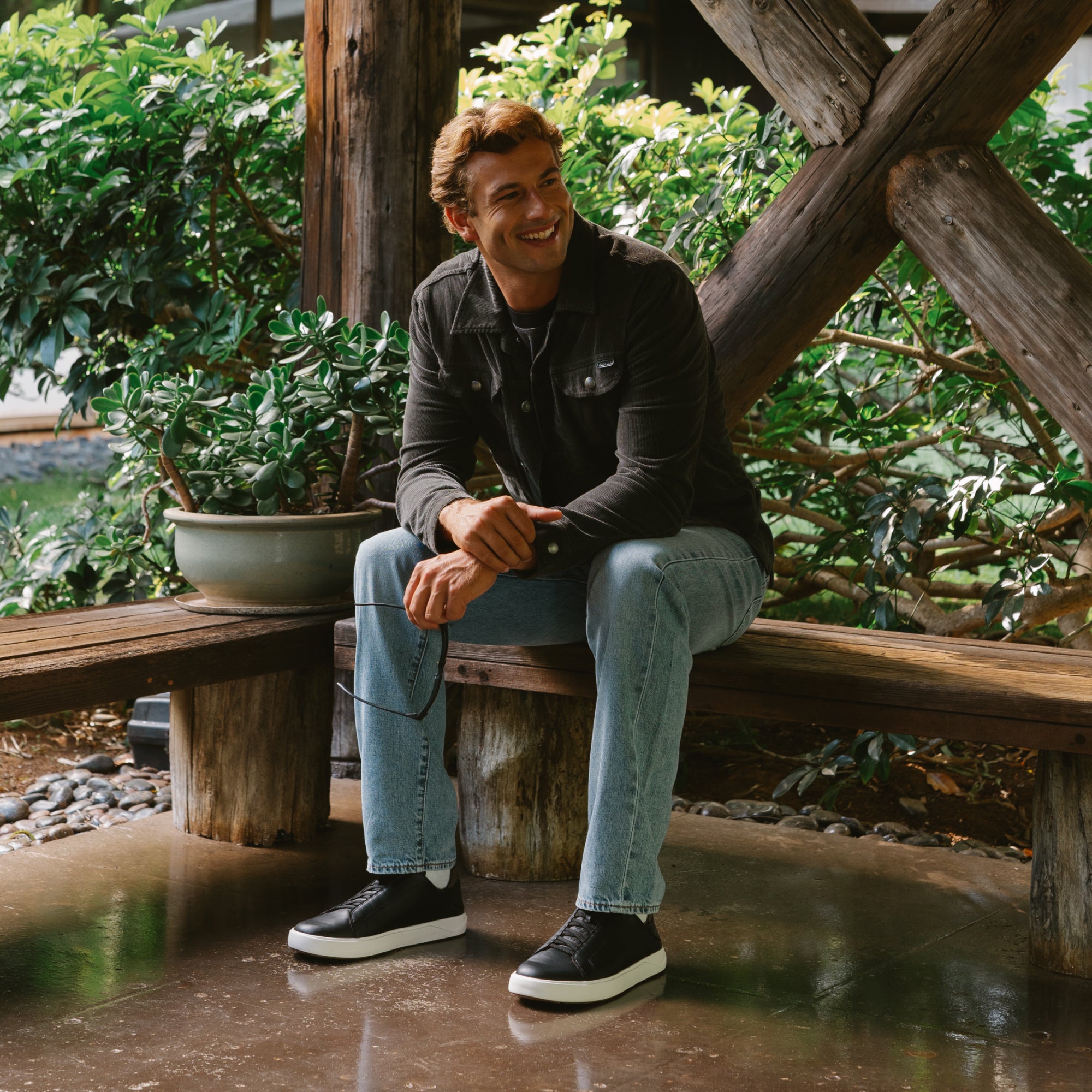 Man wearing black and dark java slip-on sneakers with white sole, seated on wooden bench in garden setting.
