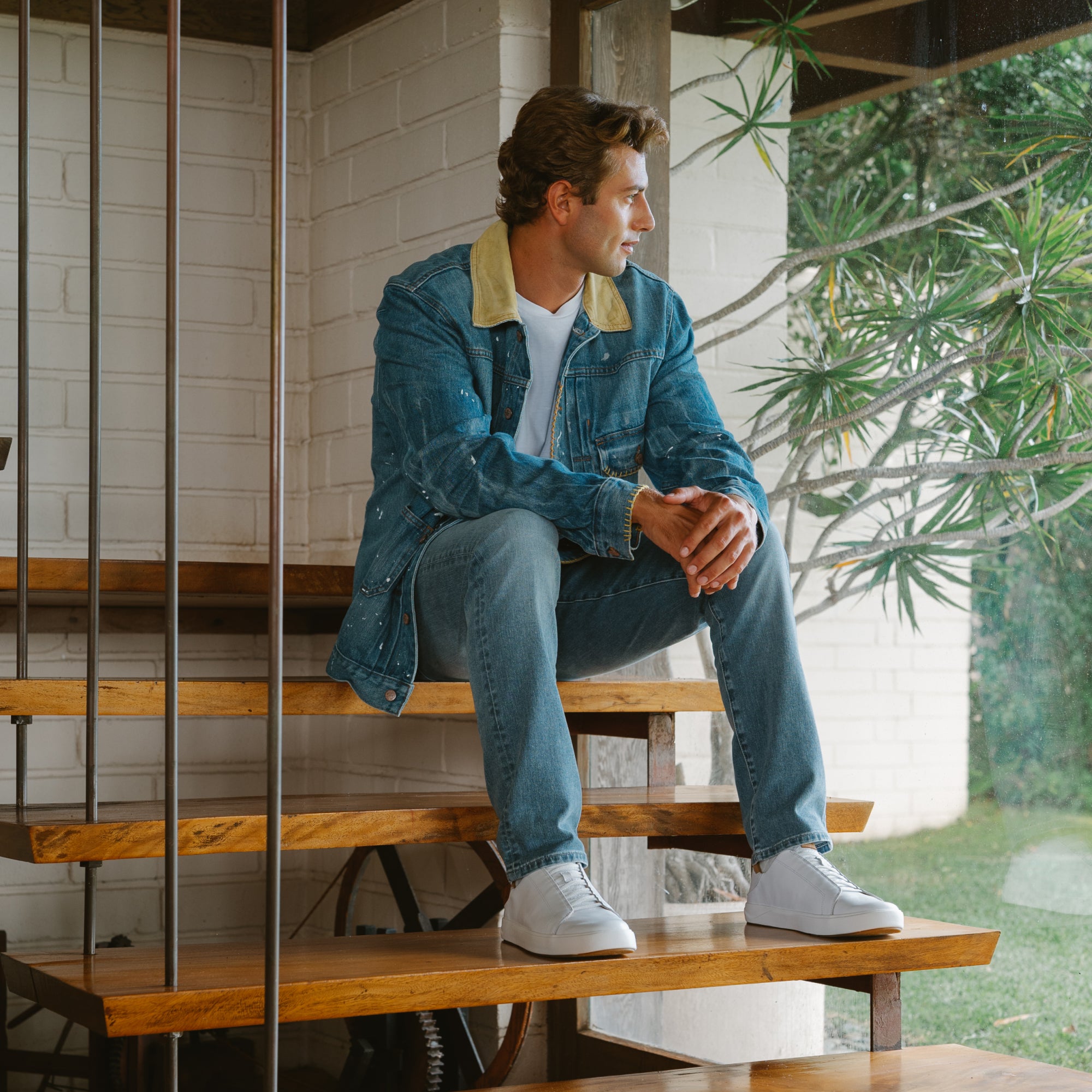 Man wearing bright white sneakers, denim jacket with yellow collar, and jeans, seated on wooden bench.