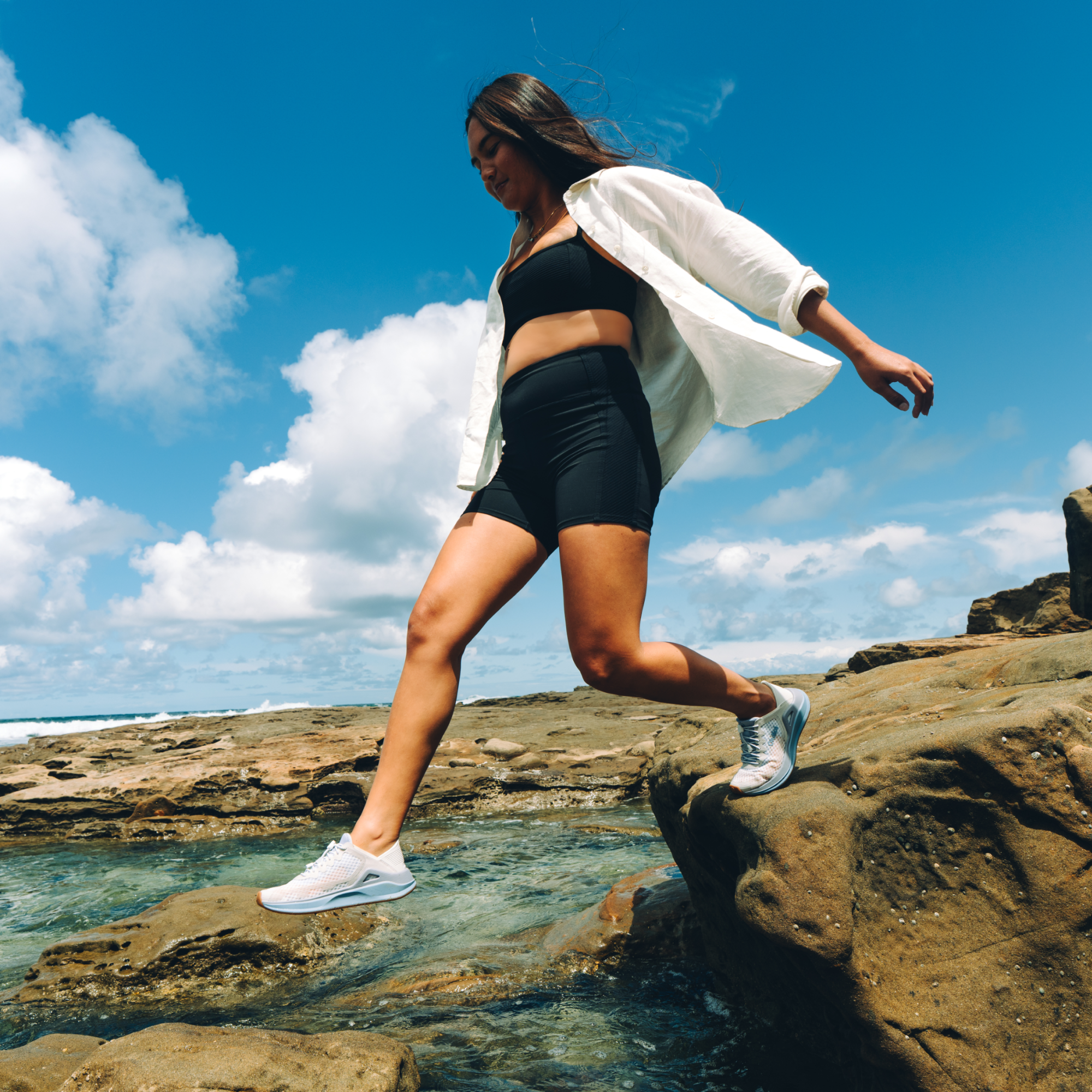 Woman wearing bright white open cardigan over black sports bra and shorts, jumping on rocky beach.