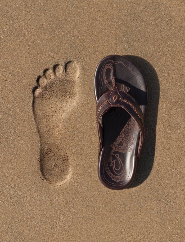 Left footprint in sand next to right Olukai sandal.